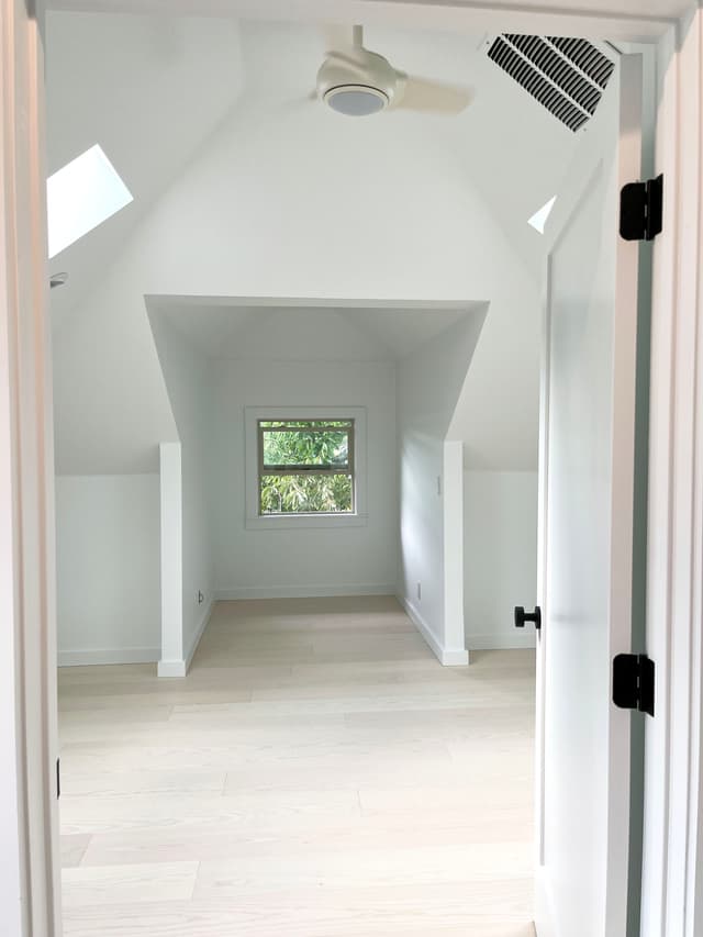 A white, brightly lit attic office with a desk in a dormer window.